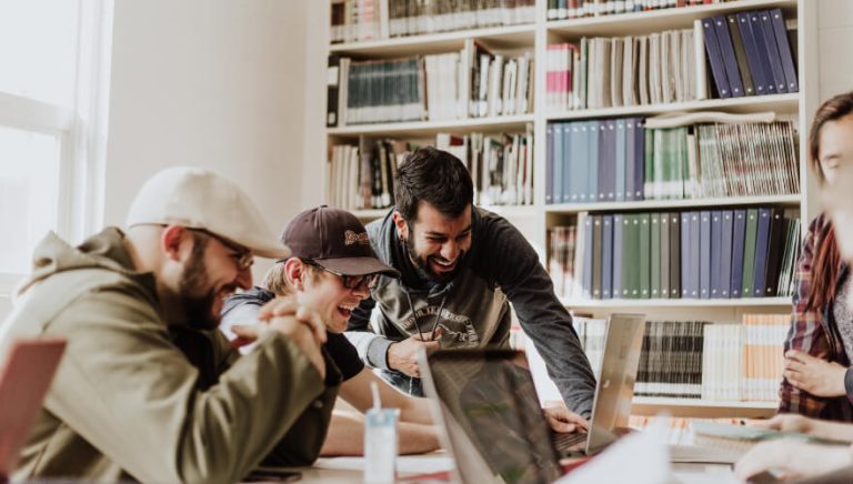 Trois hommes côte à côte qui sont dans une bibliothèque devant un ordinateur en train de rire.